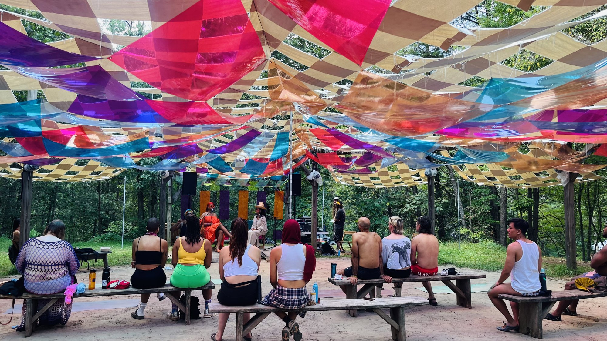 An audience on benches listening to a talk under the canopy