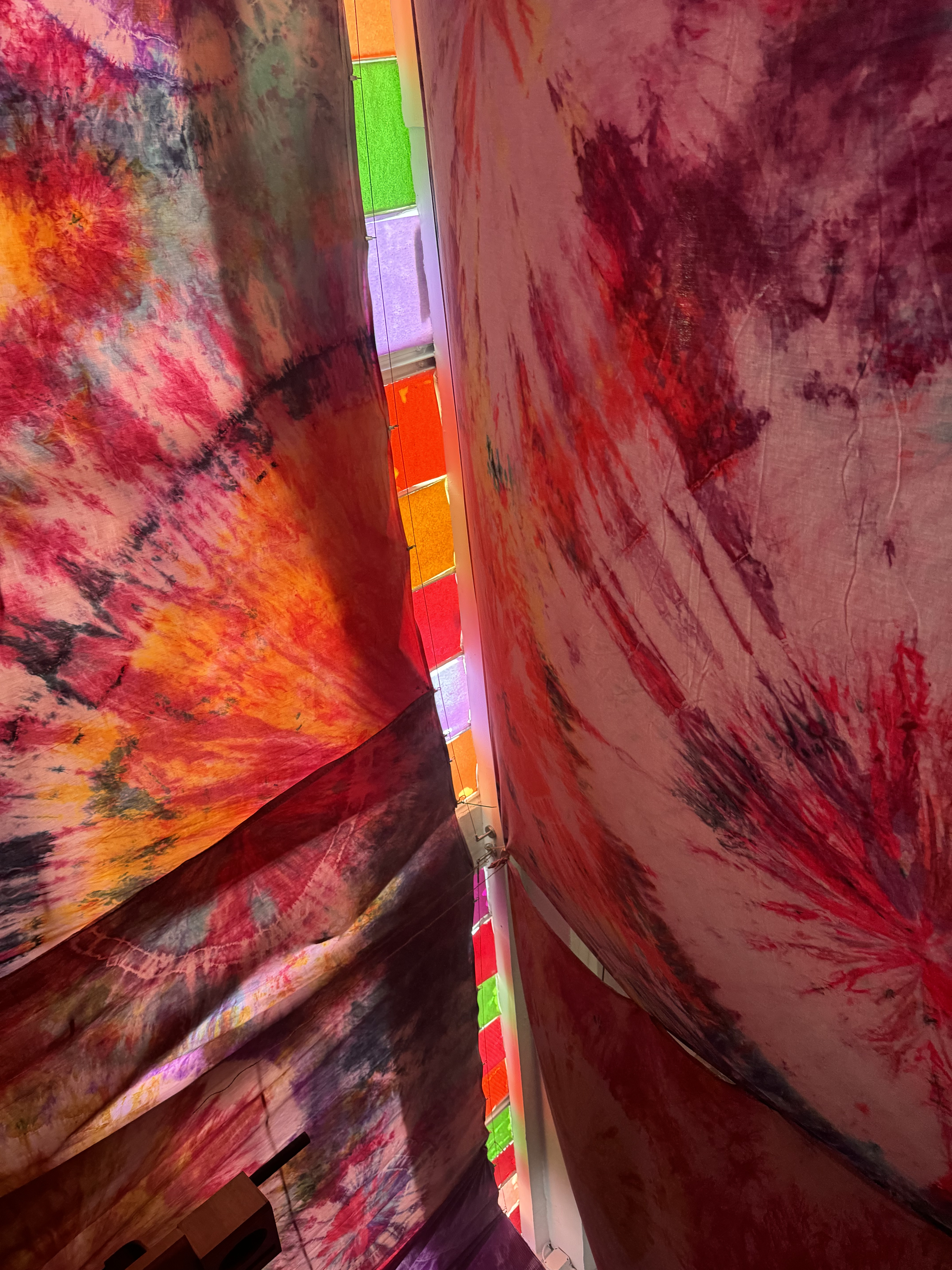 Looking up between two hanging tie-dye panels toward a strip of colored light